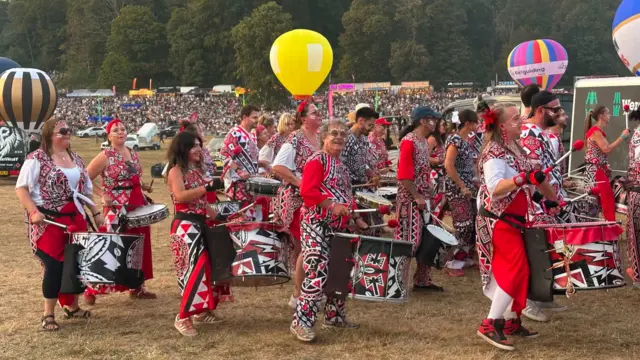 The Batala Bristol Samba-Raggae band. There is a group of people playing drums, all dressed in red, white and black.