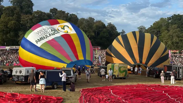 Two hot air balloons lie on the ground as they are inflated with a large crowd watching in the background