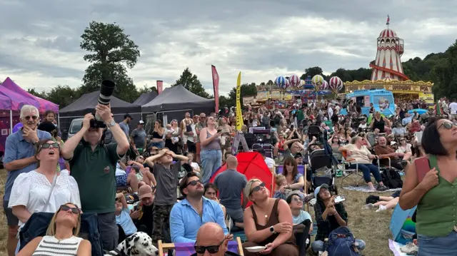 A large crowd of people looking up at the sky. Fairground rides and activities can be seen in the distance.