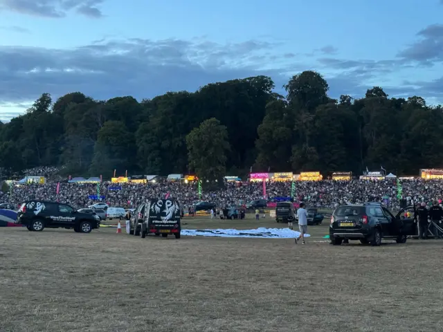 Cars and deflated hot air balloons in the middle of a field. A large crowd can be seen in the distance