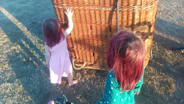 Two young girls with red hair stand with their backs to the camera, touching a balloon basket on the playing field.