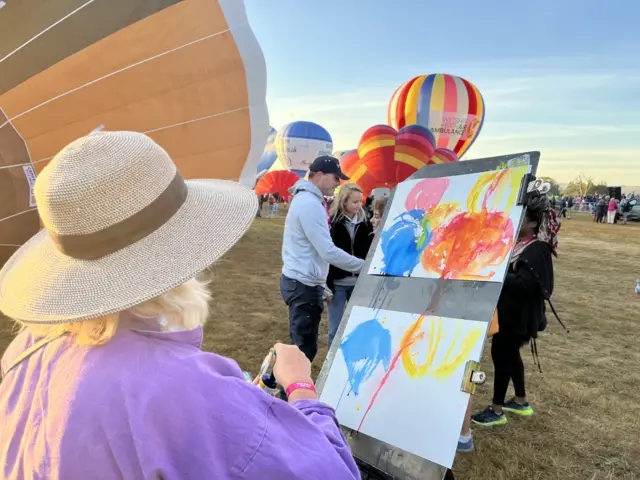 An artist wearing a wide brimmed hat and a purple shirt paints stylised balloons in different colours onto a canvas. A field of inflated balloons can be seen in front of her.