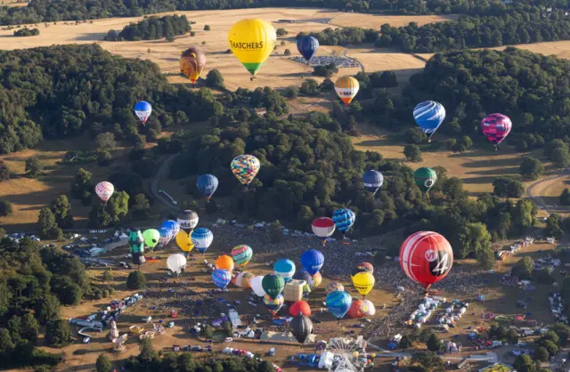 Looking down on the Bristol Balloon Fiesta site with dozens of hot air balloons taking off watched by thousands of people