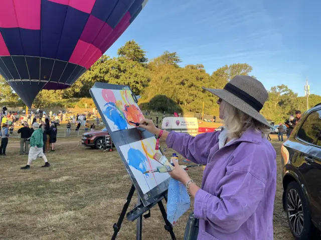 An artist wearing a wide brimmed hat and a purple shirt paints stylised balloons in different colours onto a canvas.