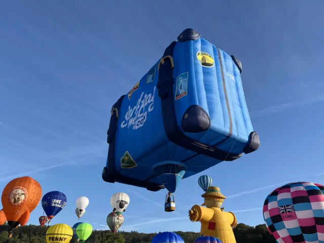 A huge, square blue balloon in the shape of a suitcase rises up from Ashton Court.