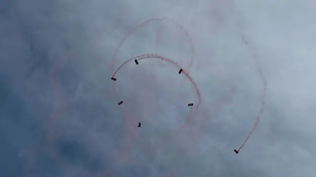 A team of RAF personnel parachuting from a plane. They are in a spiral shape and there is red smoke trailing behind them.