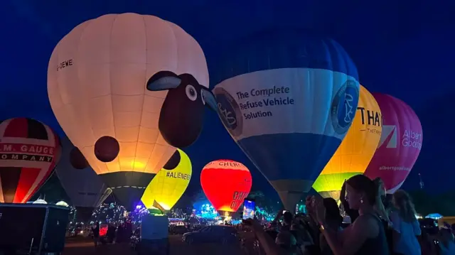 Hot air balloons glow against the night sky. They are on the ground as people watch on.