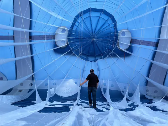 A man stands inside a blue balloon, watching it inflate.