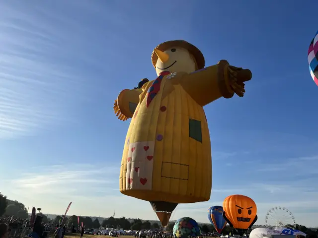 A giant yellow scarecrow balloon inflates in the field as Ashton Court as crowds look on.