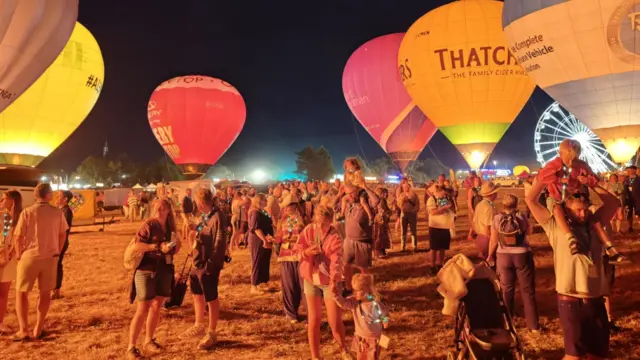 Groups of families are seen under a series of hot air balloons, with the scene illuminated by their burners