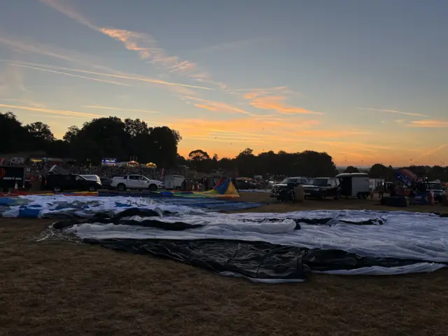 A blue and orange sunrise emerges above the busy festival site, with deflated balloons across the ground and crowds visible in the background.