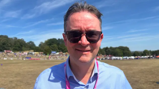 Ben Hardy. He has short dark hair and is wearing dark sunglasses, a blue shirt and a pink lanyard.