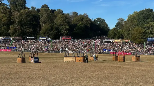 A field with multiple hot air balloon baskets in the middle of it. There is a crowd opposite.