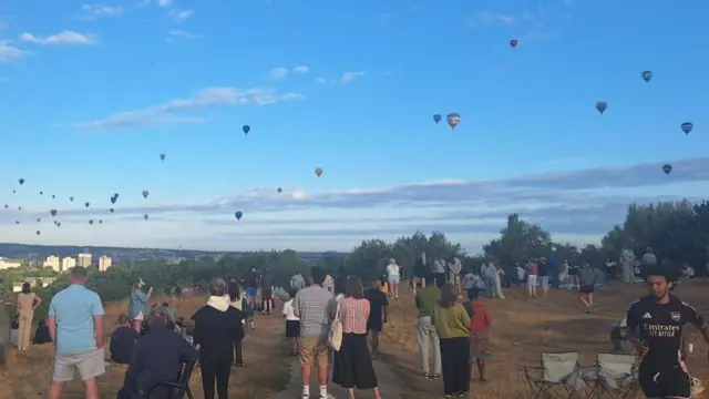 People are standing on a hill looking over a city with balloons in the sky.