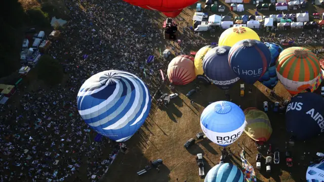 An aerial view of a mass launch at the Bristol Balloon Fiesta. Several colourful hot air balloons are being inflated next to a large bank covered with hundreds of spectators