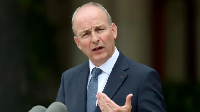 Micheál Martin speaks while wearing a navy suit and navy tie. He is standing behind a pair of microphones.