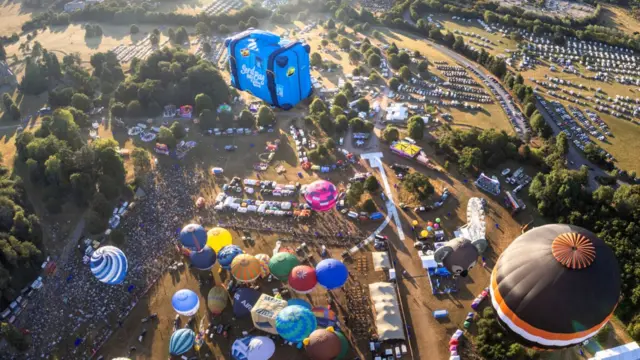 A large crowd is seen on the ground at the Bristol Balloon Fiesta as multiple hot air balloons are taking off for a mass ascent. The picture is taken from a balloon that has already taken off