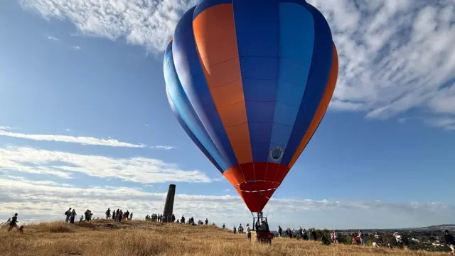 A balloon is landing on a hill with people waiting and watching.