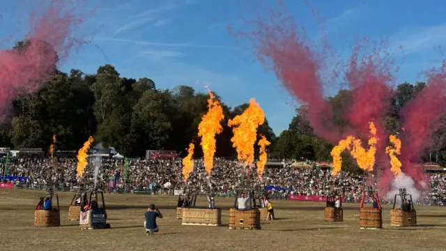 Crowds watch as fire and red smoke comes out from hot air balloon baskets