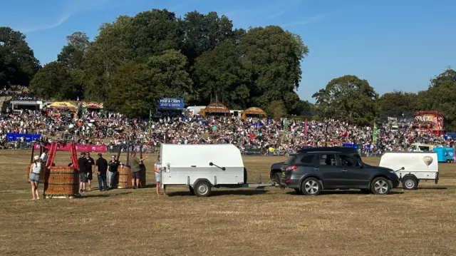A couple of cars and trailers in the middle of a field. There are also hot air balloon baskets and people tending to them.