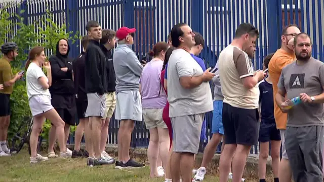 Numerous people stand outside a blue fence.
