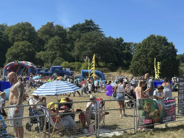 A crowd of people in a field. One group is huddled under a parasol while others are sat on the grass.