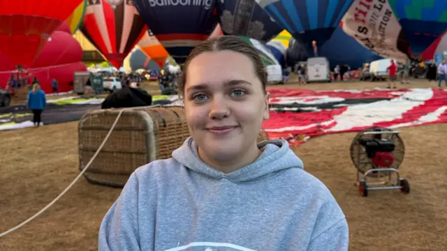 A woman in a grey hoodie is standing on a field in front of hot air balloons.