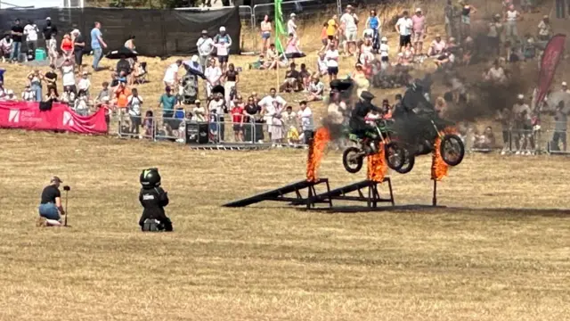A team of bikers performing a stunt show in a field. There's two riders going up a ramp and jumping through two posts that are alight.