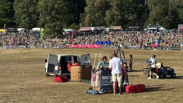 Several hot air balloon baskets in the middle of a field. Some people are tending to them.