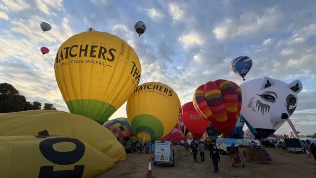 Colourful balloons are inflating on the ground while some are flying in the sky behind them.