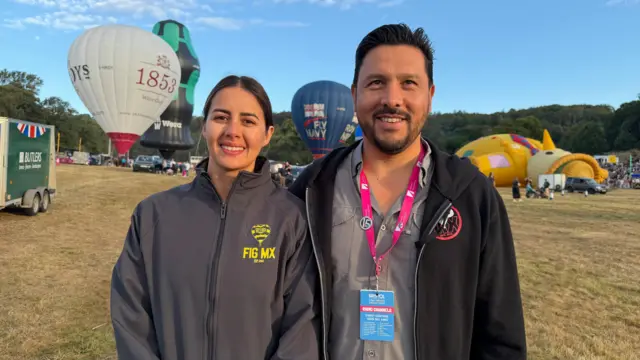 A man and a woman are standing next to each other on a field with balloons behind them.