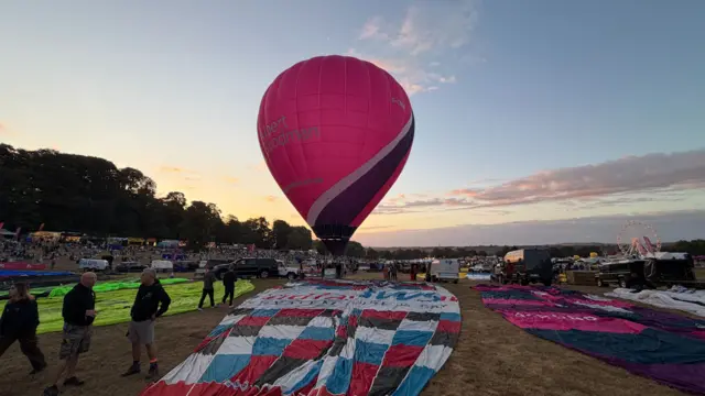 A balloon isinflated in front of a sunrise, with other balloons flat on the ground.