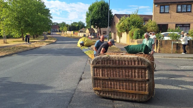 A balloon and its basket have landed on a road as people pack it back in.