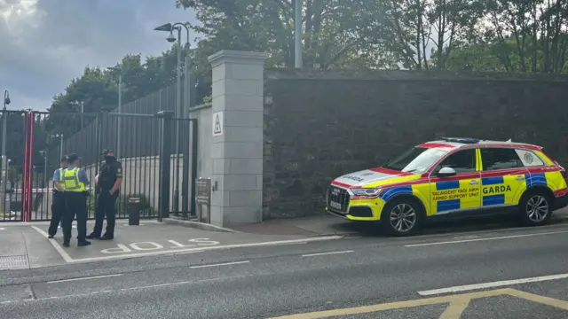 Police stand outside a gate with a police car saying "Garda" visible packed nearby.