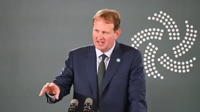 A middle-aged man with reddish blond hair mid-speech from a stage. He's standing behind a podium in a dark blue suit, white shirt and red tie, his right arm slightly raised a he gestures with his hand