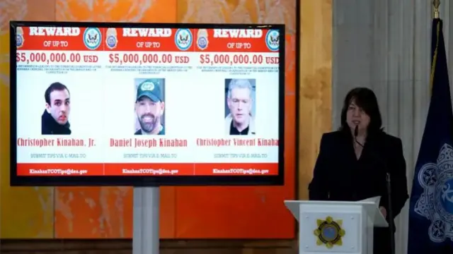 Claire Cronin speaking behind a lectern, with a screen behind her showing three wanted men