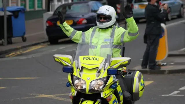 Police motorcyclist riding with both hands aloft.