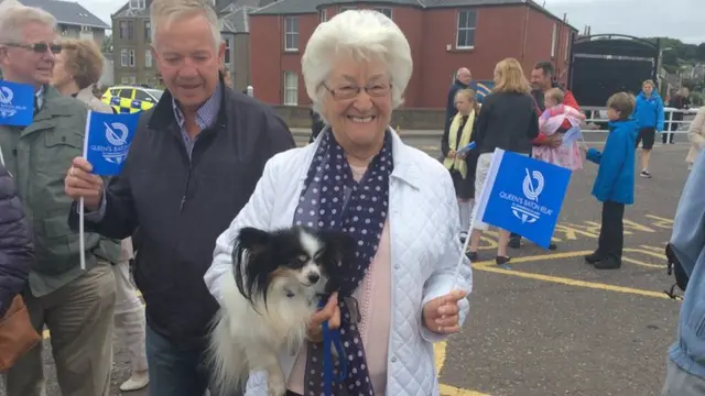 Woman holds small dog while waving Queen's Baton Relay flag