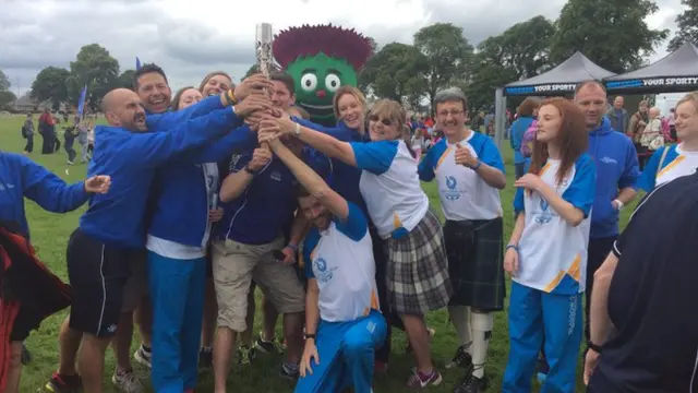 A scrum of people grasp the Queen's baton. Mascot Clyde stands at the back.