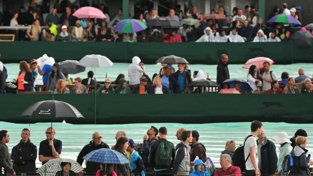 Spectators stand by the covered outer courts with umbrellas as rain suspends play