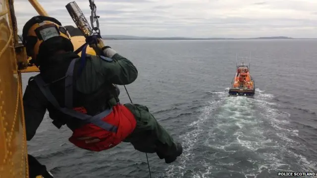 Man dangles from side of airborne helicopter holding Queen's Baton above an RNLI lifeboat.