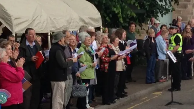 Large choir singing. A small Clyde mascot is visible in the corner of the picture, peeking into the frame.