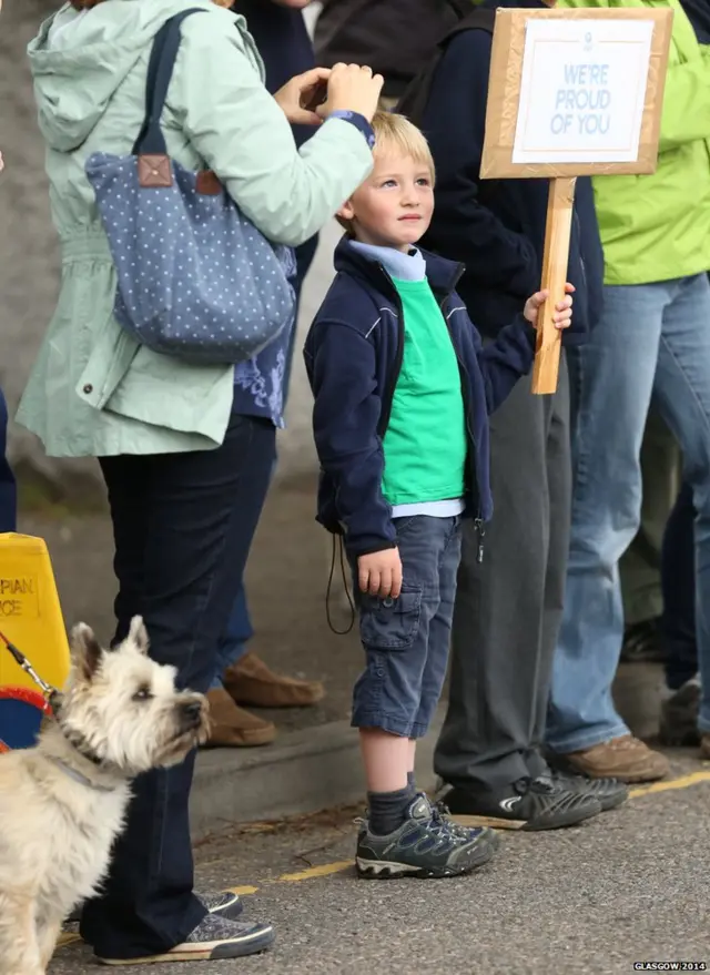 Boy holds placard saying 'We're proud of you'. Small dog beside him looks slightly menacing.