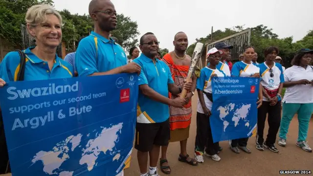 Man poses with Queen's baton as people hold banner saying "Swaziland supports our second team Argyll & Bute"