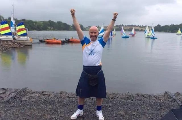 A kilted Craig Hill, arms aloft beside a loch