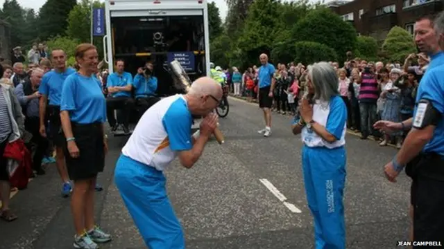 Baton bearer bows as next baton bearer presses hands together in greeting