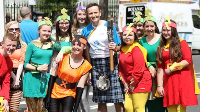 Baton bearer surrounded by people in homemade superhero costumes with headbands of dollar signs, lightning flashes, crosses and the mathematical symbol pi.