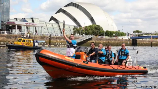 Baton bearer stands in prow of inflatable boat with Glasgow's Clyde auditorium in the background