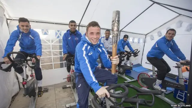 Smiling male athletes ride static bikes with foremost holding Queen's baton