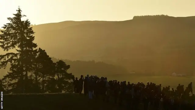 Crowds stand alongside the first fairway in early morning light